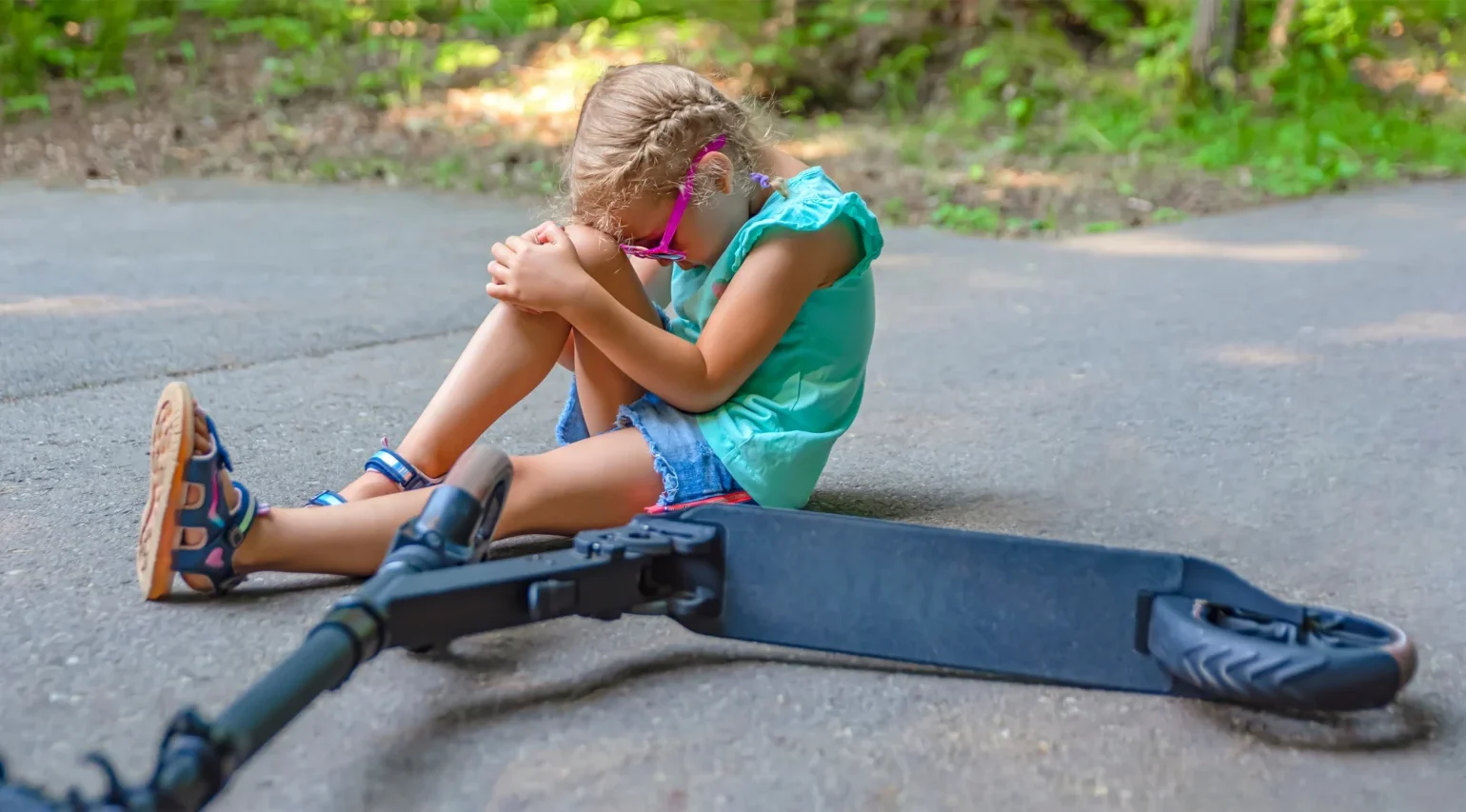 Niña pequeña en el suelo sujetándose la rodilla junto a un patinete eléctrico. Lesiones graves causadas por un patinete eléctrico defectuoso.